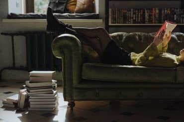 A girl lying on a sofa reading a book, with books stacked beside her